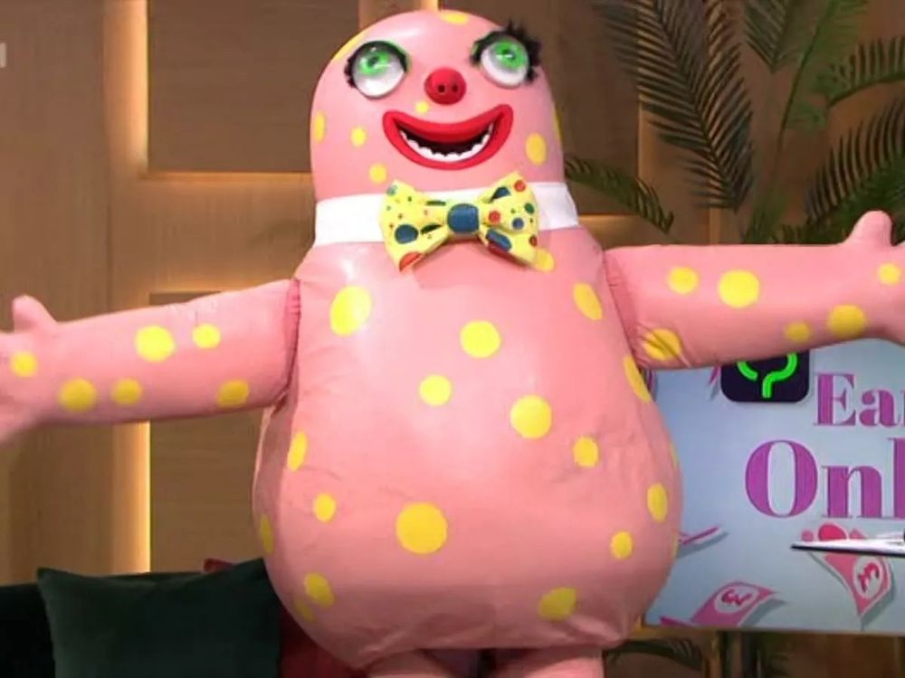 Mr Blobby. A pink skinned mascot covered in yellow spots wearing a boy, his arms stretched wide 