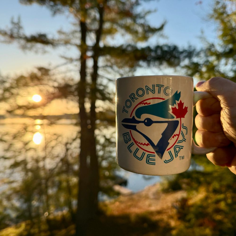 A coffee mug featuring a vintage logo of the Toronto Blue Jays baseball club being held up against a backdrop of sunrise over a northern Ontario lake. 