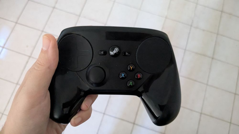 My hand holding one of the last-gen Steam controllers that you could get for like $4 before they stopped making them (I guess because they were getting patent trolled over the back paddles) over my kitchen floor which is also my living room floor they're the same room I don't need a fancy apartment man what you want me to pay for more walls I don't even want or something??? There were multiple years this was the only controller I had in the house and I wasn't complaining!!! Nowadays I mostly just use it as a remote for when I'm watching TV from my couch but when friends are over I give them the normal xbone controller and recall my freak nature.