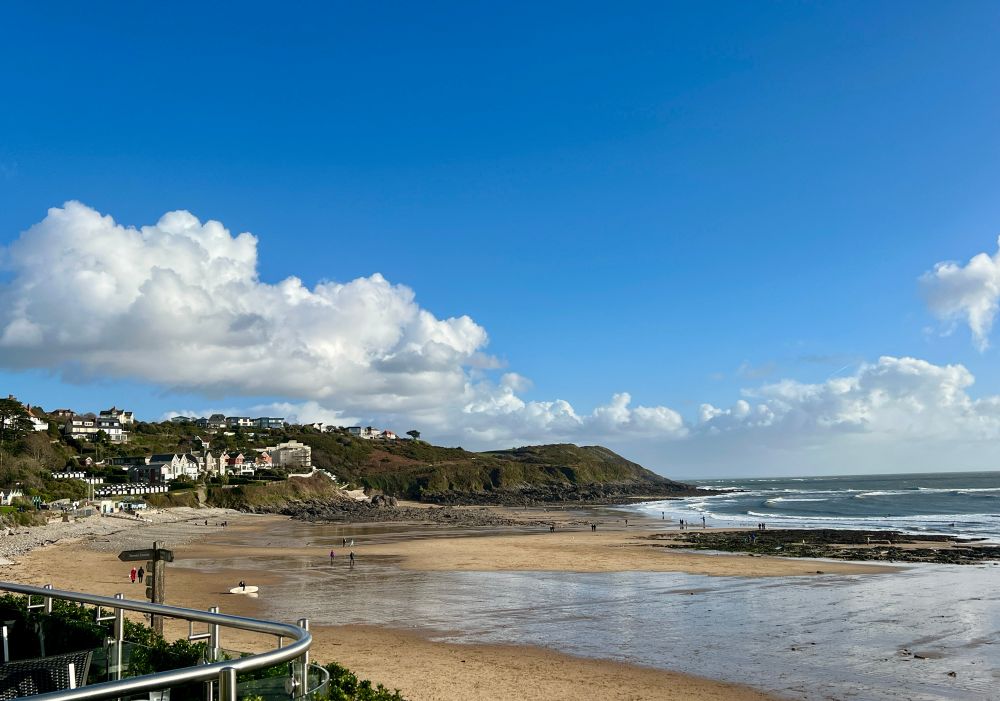 View of a low-tide sandy bay with beach wet from freshwater streams and rockpools in the distance. Puffy white clouds low in a sunny blue sky. 