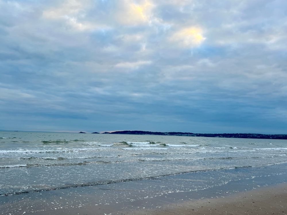 View of the blue-green bay with gently breaking low waves and a cloud-mottled sky with bright patches. 