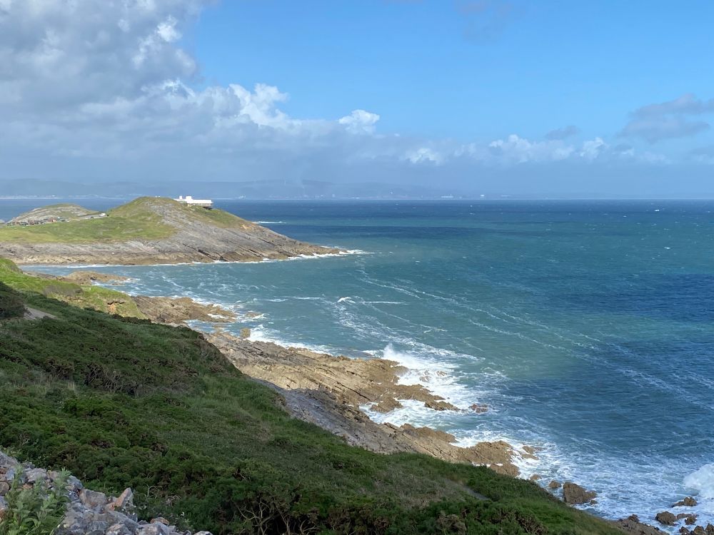 View of the South Gower coast to Mumbles Head and the old coastguard station shining white in the sun. Blue sea with white surf breaking on the rocks and mostly blue sky with cloud in the distance. 