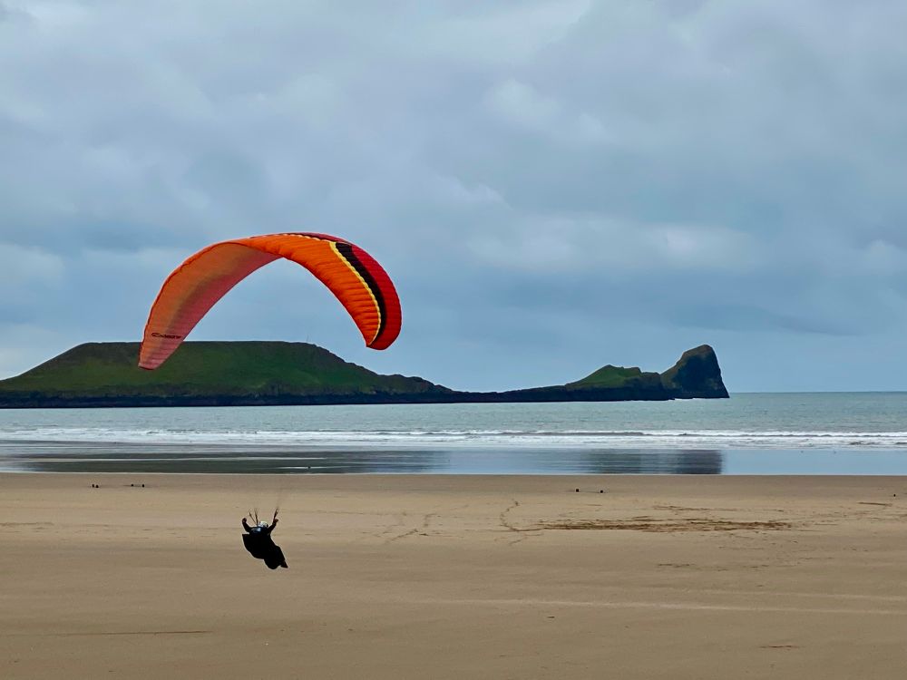 A paraglider with big orange parachute coming in to land low over the golden sand of Rhossili Bay. Worm's Head is laid out dark on the horizon. 