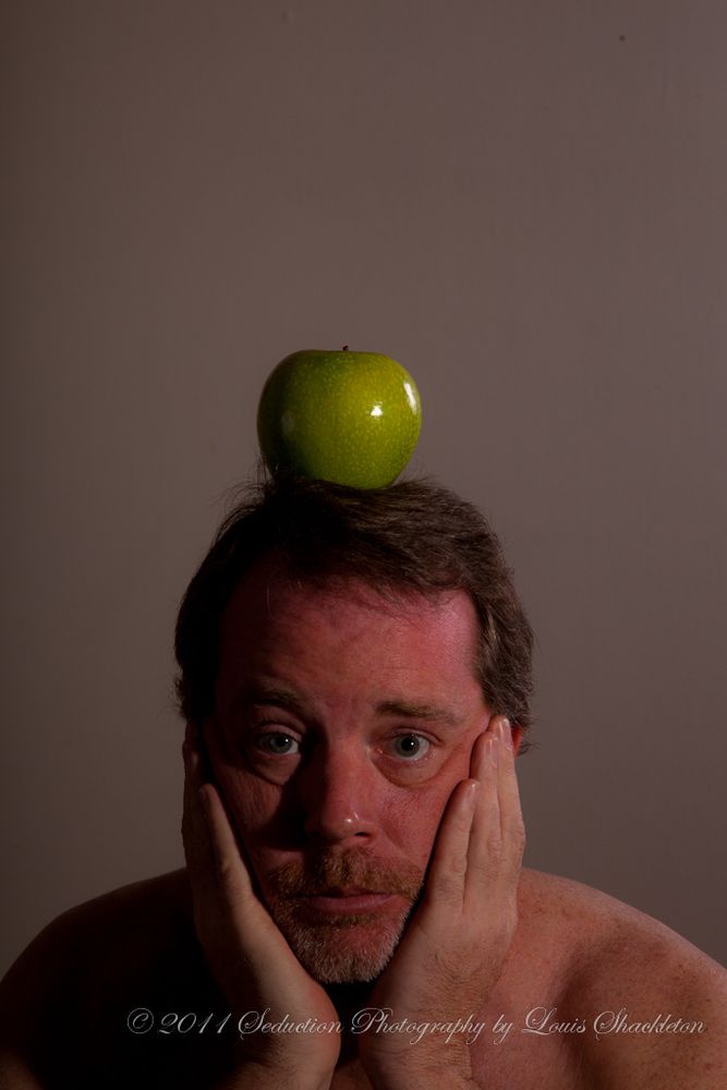 “Ceci n’est pas une pomme”, self portrait. Best of show, 2011 All Student Art Show, UNCW. A brown-haired white man with blue eyes and a goatee faces the camera chin in palms, with a green apple perched on his head, against a plain gray background in an homage to René Magritte, Belgian Surrealist painter from the first half of the 20th century. 
