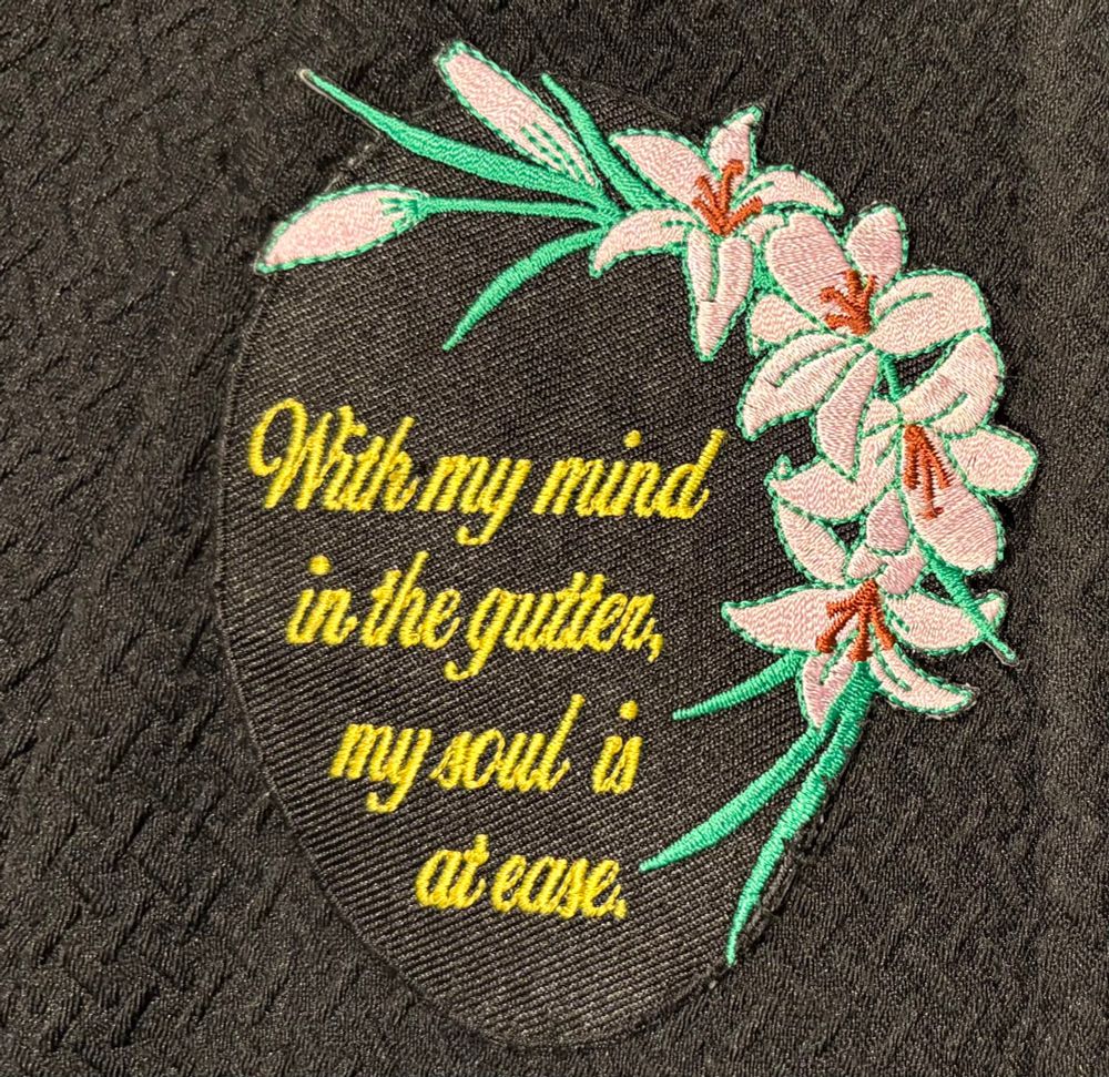 A photo of an oval shaped patch sewn into a black jacket. The patch is black. Yellow cursive script reads, “With my mind in the gutter, my soul is at ease.” Pink lilies and leaves border about half the edge.
