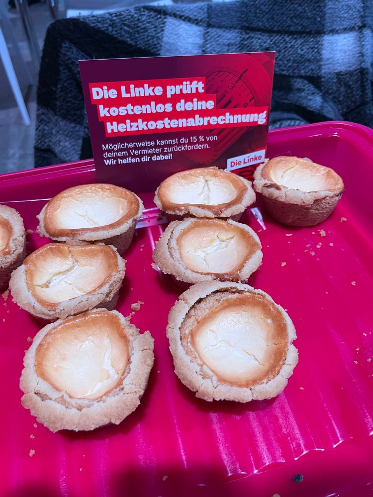 Das Bild zeigt kleine Käsekuchen-Muffins auf einem roten Tablett. Im Hintergrund steht ein Schild der Partei “Die Linke” mit dem Text: „Die Linke prüft kostenlos deine Heizkostenabrechnung. Möglicherweise kannst du 15 % von deinem Vermieter zurückfordern. Wir helfen dir dabei!“