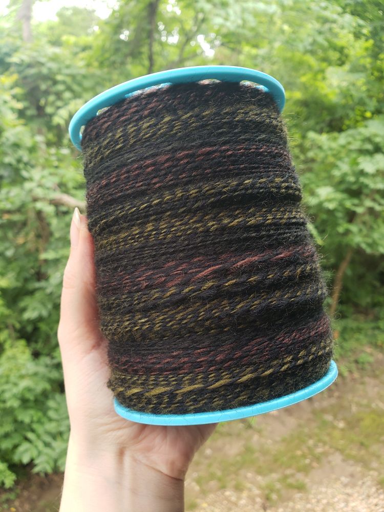 A bobbin full of handspun yarn. The yarn is a swampy brown/green/dark yellow mix plied with solid black.