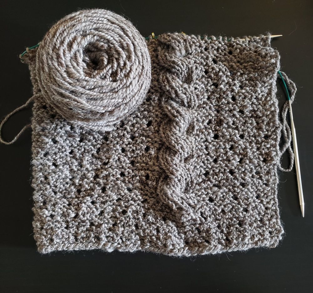 A knit project in progress: a rectangular wrap with eyelets and a large cable detail on one side, knit in a rustic, oatmeal colored, handspun yarn.