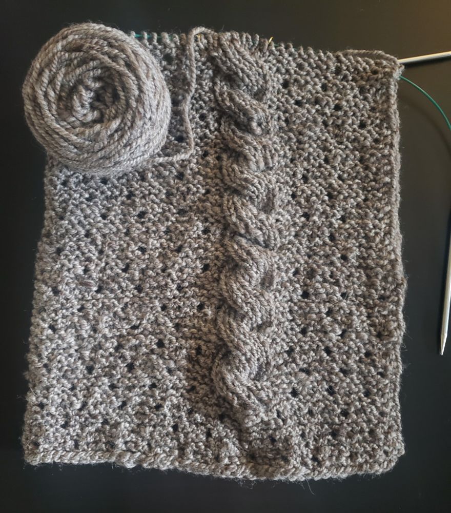 The same knit project in progress: a rectangular wrap with eyelets and a large cable detail on one side, knit in a rustic, oatmeal colored, handspun yarn. The project is a couple of inches longer in this photo.