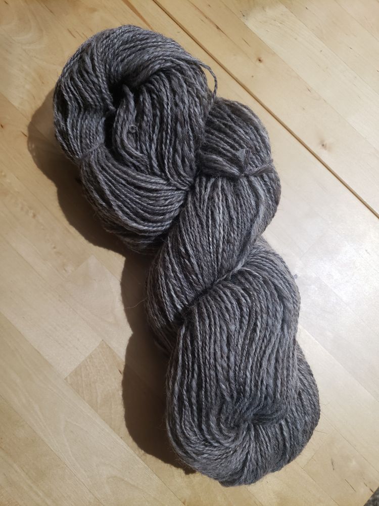 A large, loosely twisted skein of the same yarn, which looks much more grey, laying on a table in artificial light.