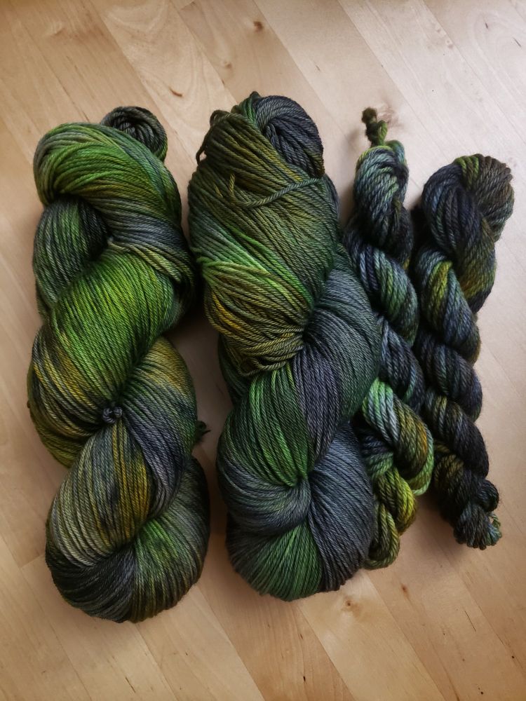 Two full size skeins and two mini skeins of yarn. They are all dyed in shades of green, blueish grey, black, yellow, and some brown.
