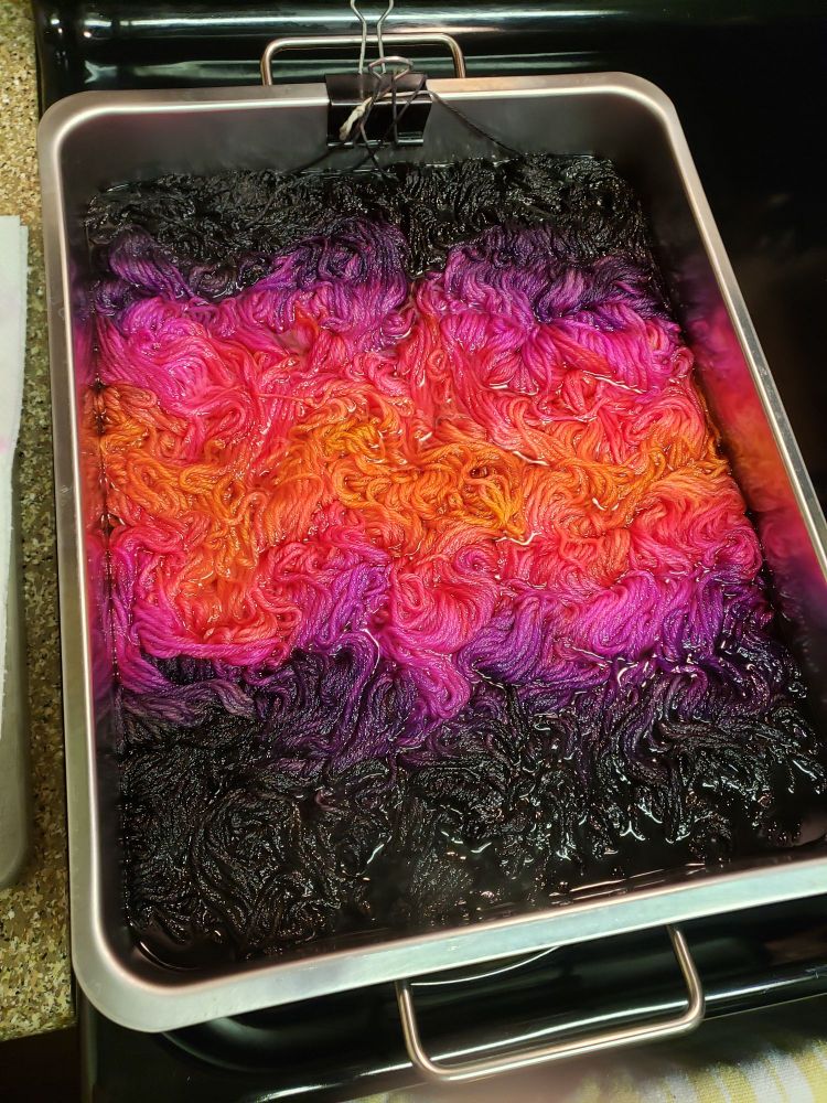 A large rectangular pan on a stove. In the pan, two skeins of yarn in the process of being dyed a vibrant sunset gradient: black, purple, fuchsia, orange, fuchsia, purple, black.
