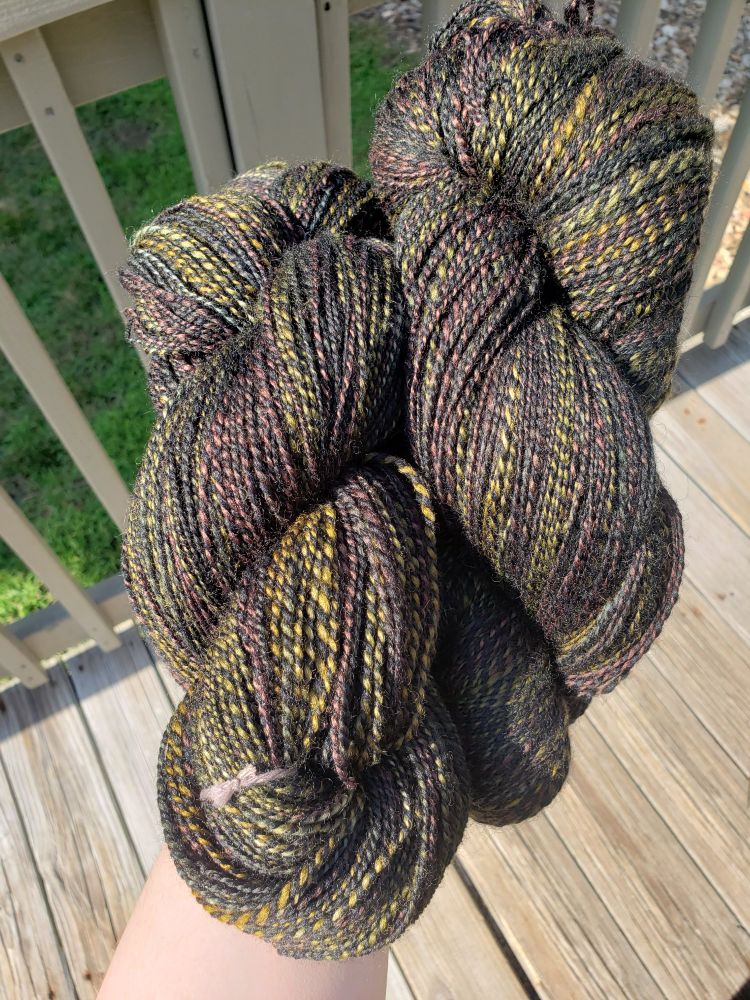 Two large hanks of two-ply handspun yarn, one ply is solid black, one is various shades of green and brown. Photographed in direct natural light.