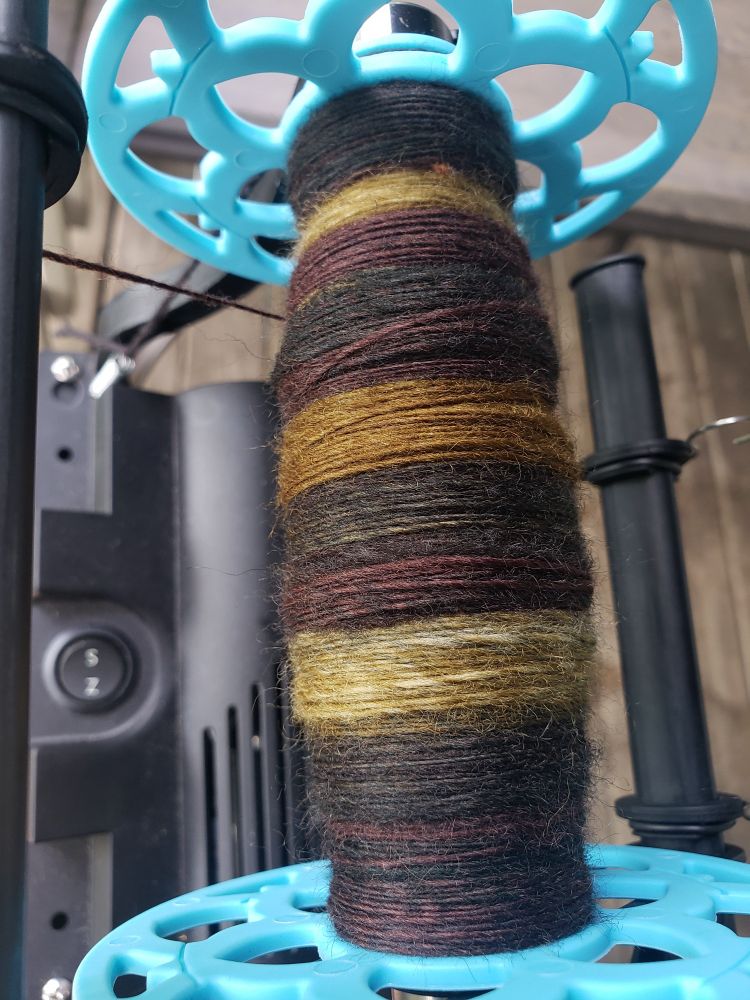 A handspun single on an e-spinner bobbin, in the sunlight. The yarn is a deep warm brown, dark forest green, and muted golden color. The colorway name, peat bog, is accurate.