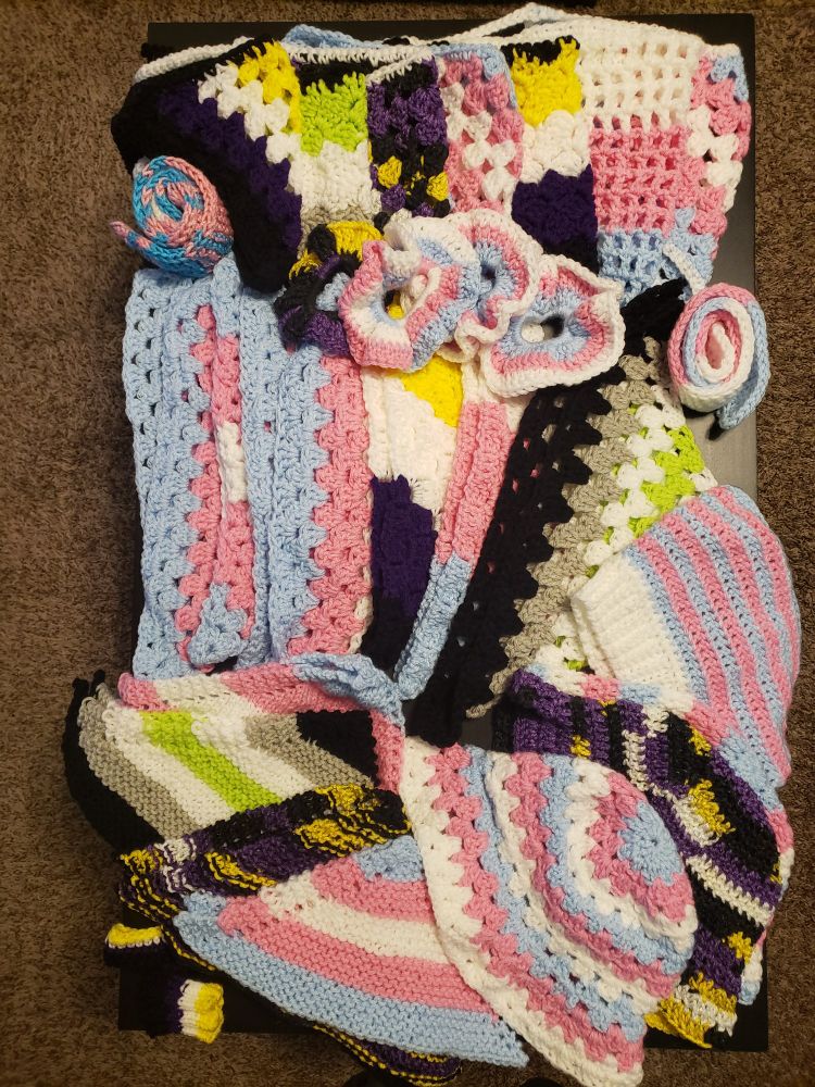 A coffee table covered in knit and crochet bandanas, hats, scrunchies, etc, all in trans, non-binary, and agender pride flag colors 