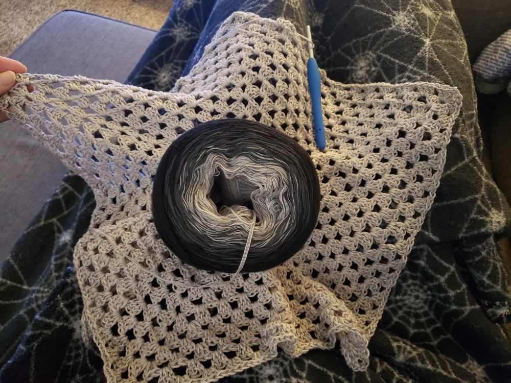 A crochet project in progress, which is the beginning of a granny hexagon wrap cardigan. The yarn is a gradient cake from light grey to black.