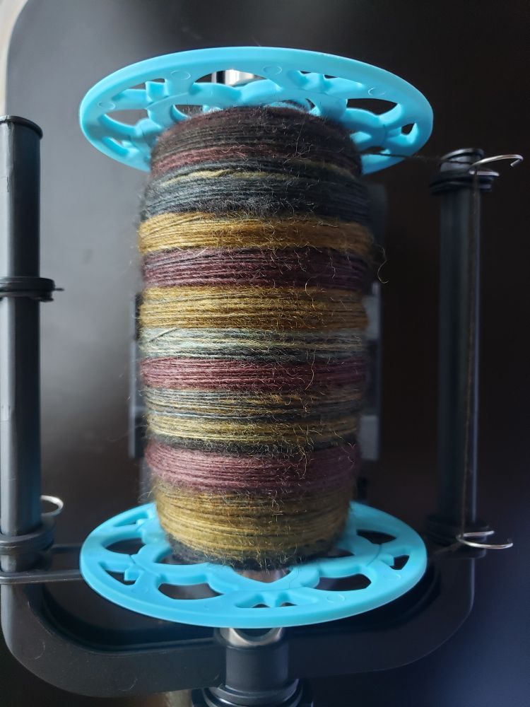 Handspun yarn on an e-spinner bobbin. The yarn is in shades of brown, green, and a dark golden yellow.