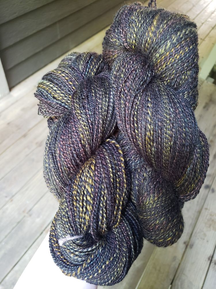 Two large hanks of two-ply handspun yarn, one ply is solid black, one is various shades of green and brown. Photographed in the shade.