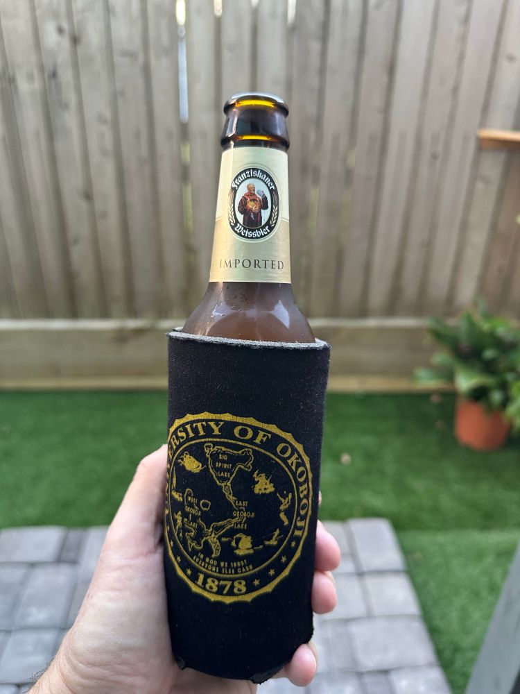 A photo of a bottle of Franziskaner Weissbier in a koozie from the University of Okoboji. 

The “university” is fictional, being dreamed up by a couple of entrepreneurs back in the 1970’s. They started a clothing shop in the town of Okoboji, Iowa and thought it’d be fun to print t-shirts with the name. 