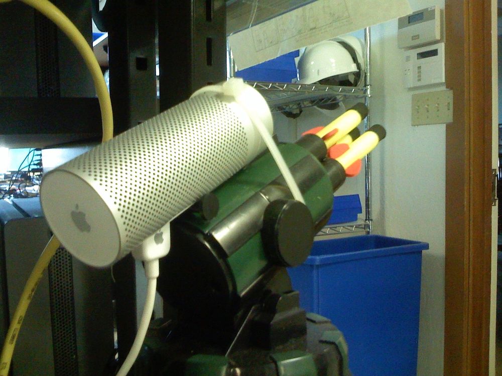 A silver cylindrical Apple iSight webcam is strapped with a zip tie to the barrel of a toy dart gun. The toy gun is green and black with several foam darts loaded at the front. The setup is on a shelf in a room with visible hard hats, bins, and network cables in the background.