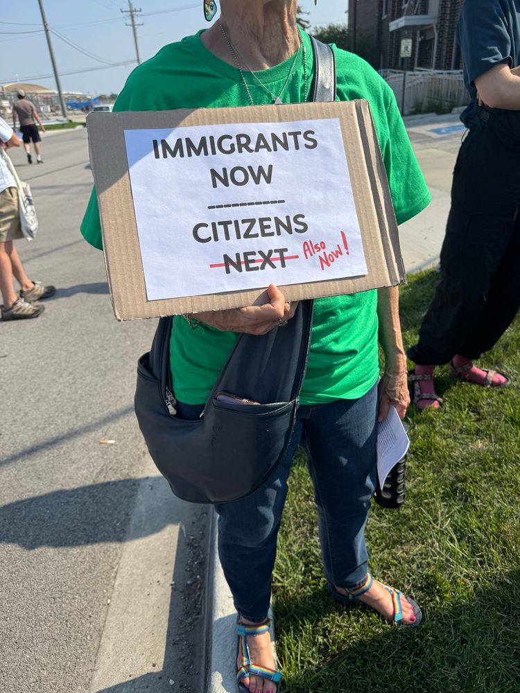 A woman holds a sign reading “IMMIGRANTS NOW / CITIZENS NEXT” but “NEXT” is crossed out and edited with red marker to say “Also Now!”