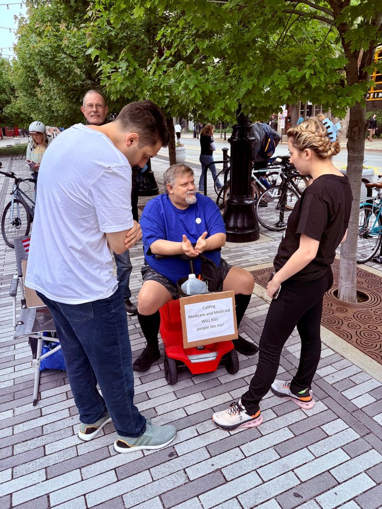 Ben and I talk to a man in a mobility aid with a sign reading “Cutting Medicare and Medicaid WILL KILL people like me!”