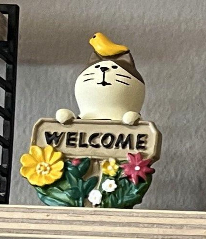 Photo of little statuette of white cat with brown ears holding a sign that says “welcome,” which is framed with yellow, red, and white flowers. A yellow bird sits on the cat’s head.