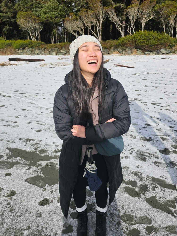 Im a Chinese woman with long black hair. I am wearing a grey knit har, long puffer jacket and leggings. I am standing on the beach, there is snow on the ground. I am standing in the sun. 
