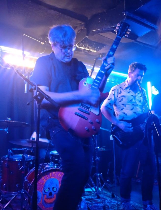 Matt Stevens mid performance with The Fierce & The Dead at Ramsgate Music Hall. Everything is bathed in blue light, and Matt is showing how he's professional enough to pull a huge chord out of the bag without pulling a face like he's followed through. The back of the shot shows bassist Kev in a now infamous polyester shirt.