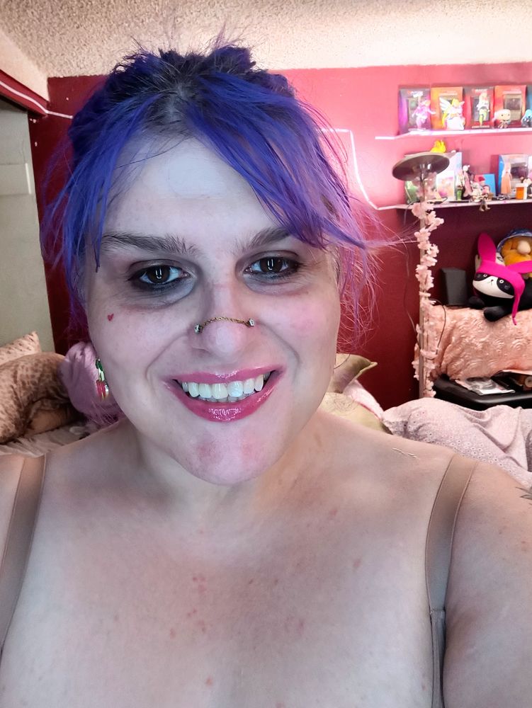 Cherry with her violet hair. A smokey eye look. She is wearing pink lip gloss and flat dangling pink cherry fruit earrings. She smiles showing teeth.