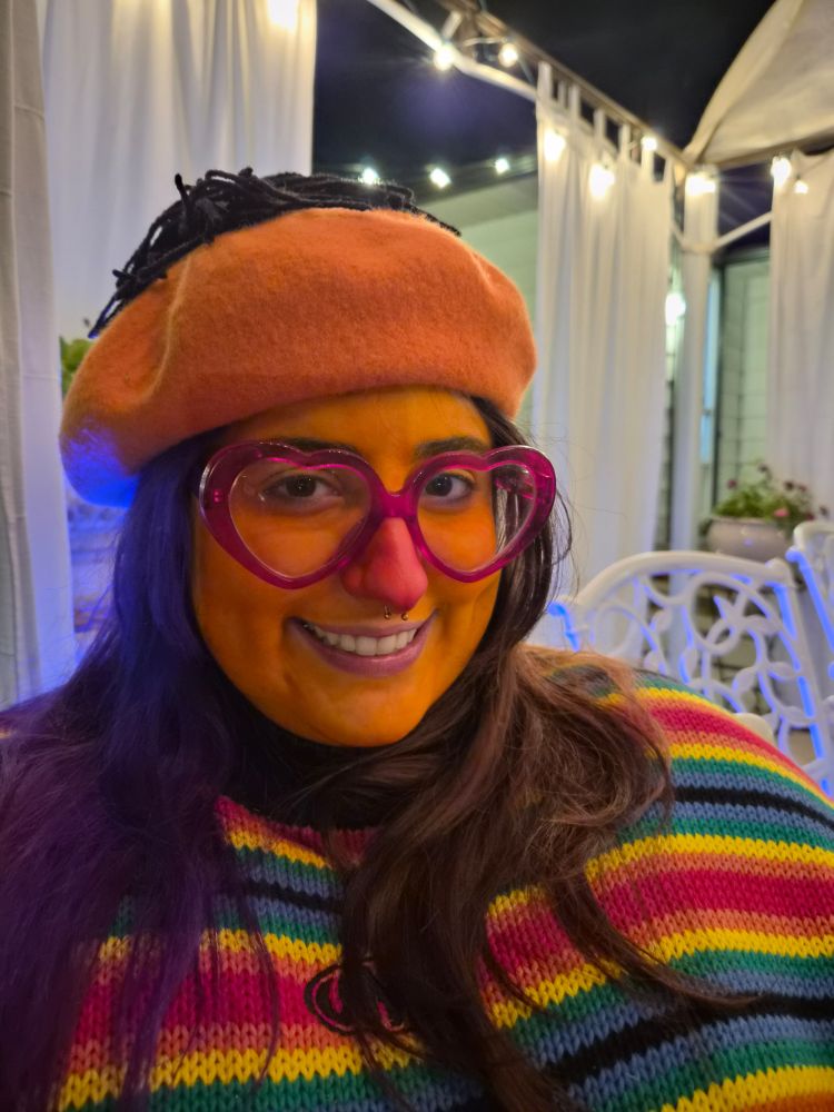 Woman dressed like Ernie from sesame street. She's wearing an orange beret with a black pompom on top, orange face paint, red nose, and striped sweater. 