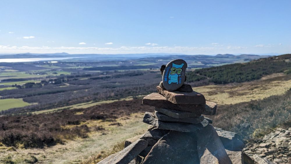 Backlit by sunshine, a small painted stone of a standing, and slightly morose looking, korok rests on top of a cairn. In the distance the hills and fields sprawl out, with the Tay cutting through them.