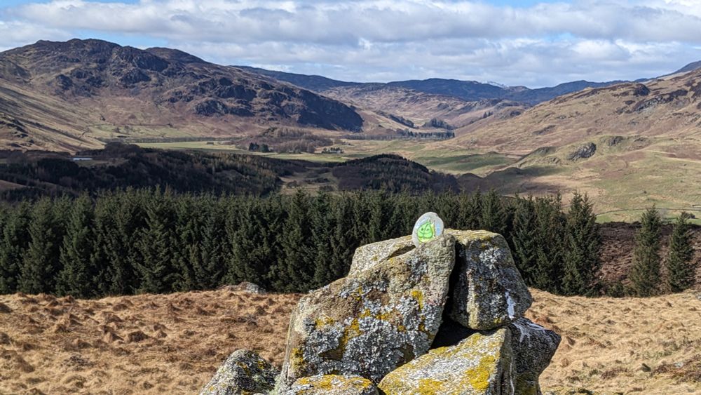 A small painted stone of a Korok with a green heart shaped leaf face, rests atop a lichen covered cairn, whilst the Scottish Highlands behind are dappled in sunlight. 