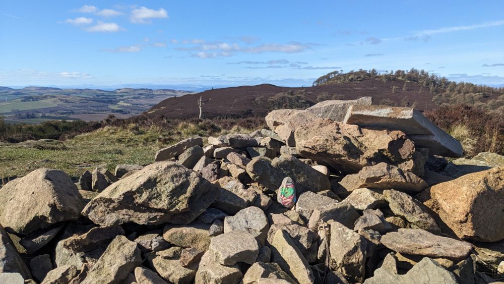 A small painted stone of korok sits upon a large cairn under a beautiful blue sky. Views of the Sidlaws and Grampians sprawl out in the background.