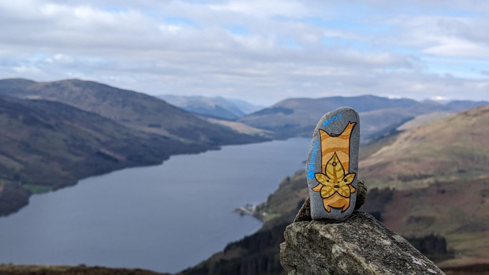 A painted rock of a Korok with an autumn coloured maple leaf face stands tall on top of a cairn, whilst the sun dappled hills around Loch Earn stretch off into the distance to some snow clad peaks. 