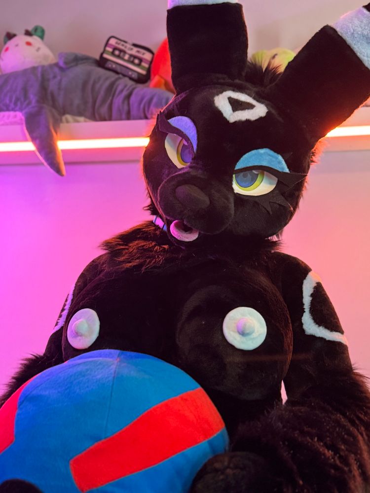A photo of a shiny female Umbreon fursuiter holding a giant plush great ball under her large boobs and light blue nipples. In the background there is a shelf full of assorted plushies including a Blahaj. The lighting is orange, purple, and blue somewhat similar to the “bisexual lighting” colors.