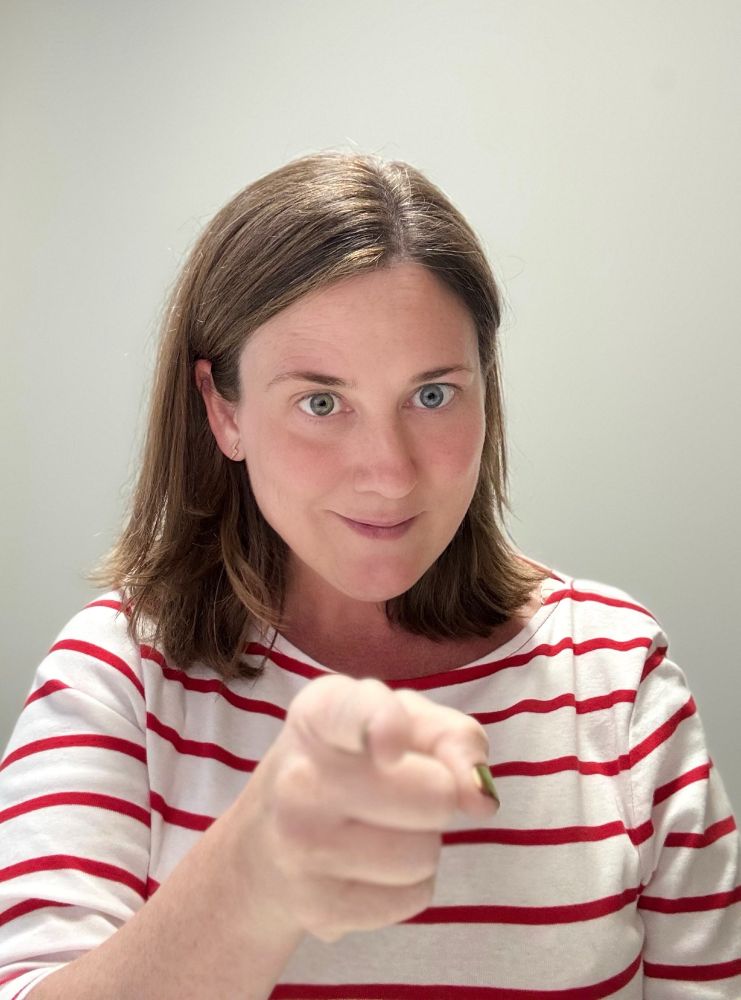 A woman with brown hair (me) in a red and white striped top, looking directly into the camera and pointing.