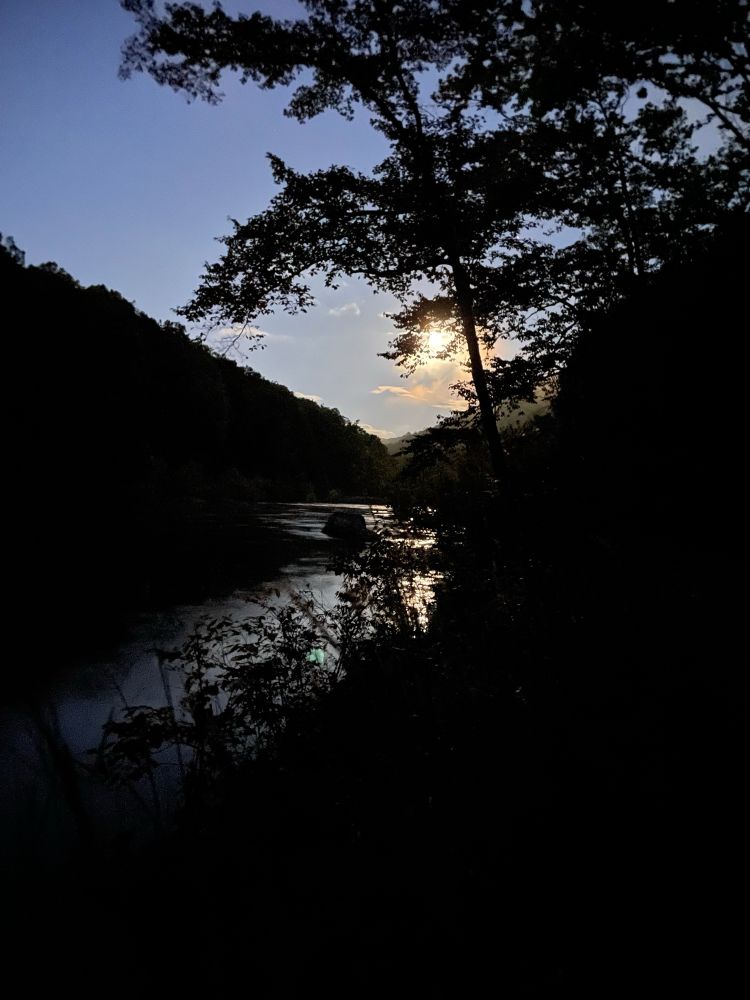 A river flows through a forested area at dusk, with the sun partially obscured by trees and its reflection visible on the water.