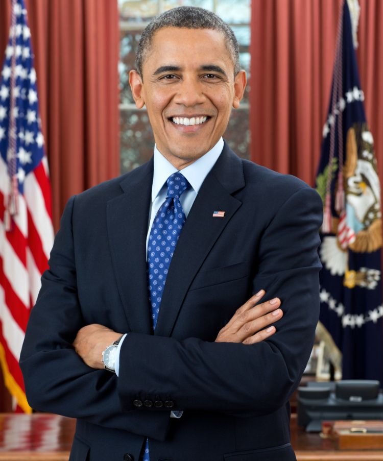 President Barack Obama with a smile and arms crossed and the flag behind him. He is wearing a blue suit and a blue tie with stars or white dots. 