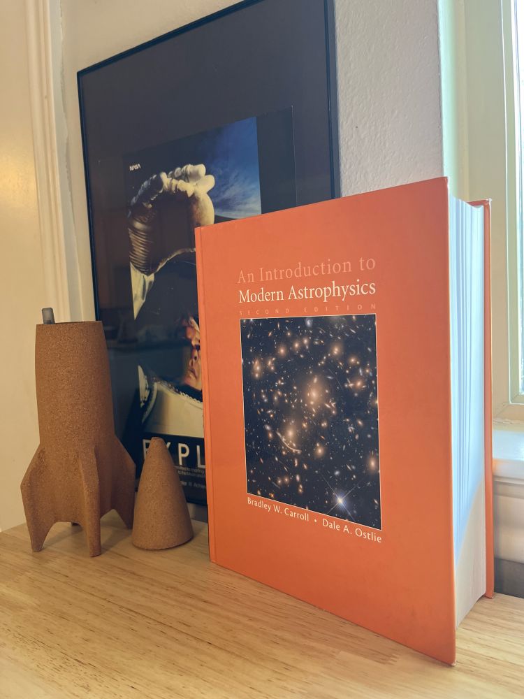 The “big orange book” of astrophysics — An Introduction to Modern Astrophysics btw Carroll and Ostlie — stood upright on a wooden desk, in front of a poster of an astronaut grabbing the moon, and next to a rocket-shaped pen-holder. 