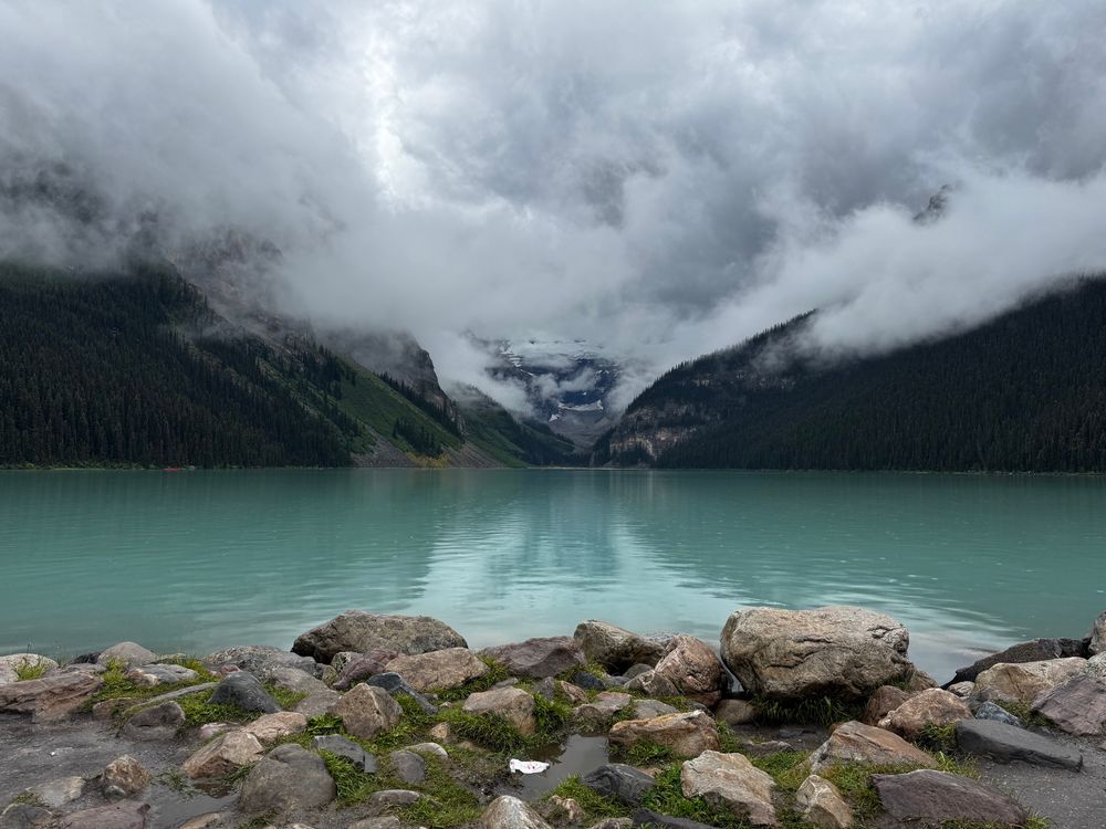 Clouds over a neon blue lake with mossy rocks in the foreground.