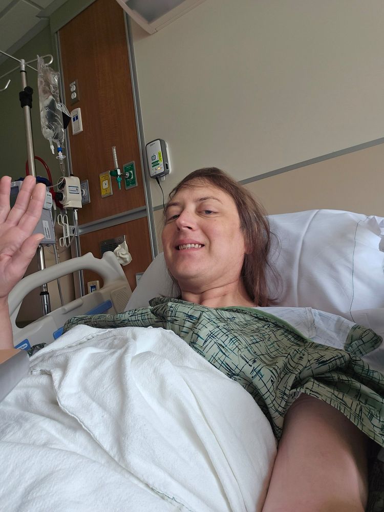 Me lying in a hospital bed looking groggy af whilsy waving with IV bag et al behind me