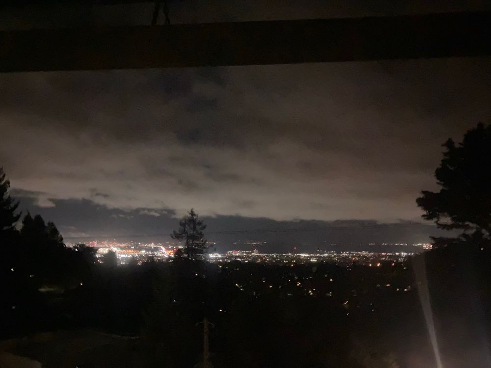 early and I mean early a.m. view of the Bay area from the Oakland Hills looking west to San Francisco.  A few lights pepper the black silhouette of the trees and hills in the foreground, giving in to brillyant lights of the bridges, roads and SF beyond.  Low clouds cover the sky and appear cottony light gray