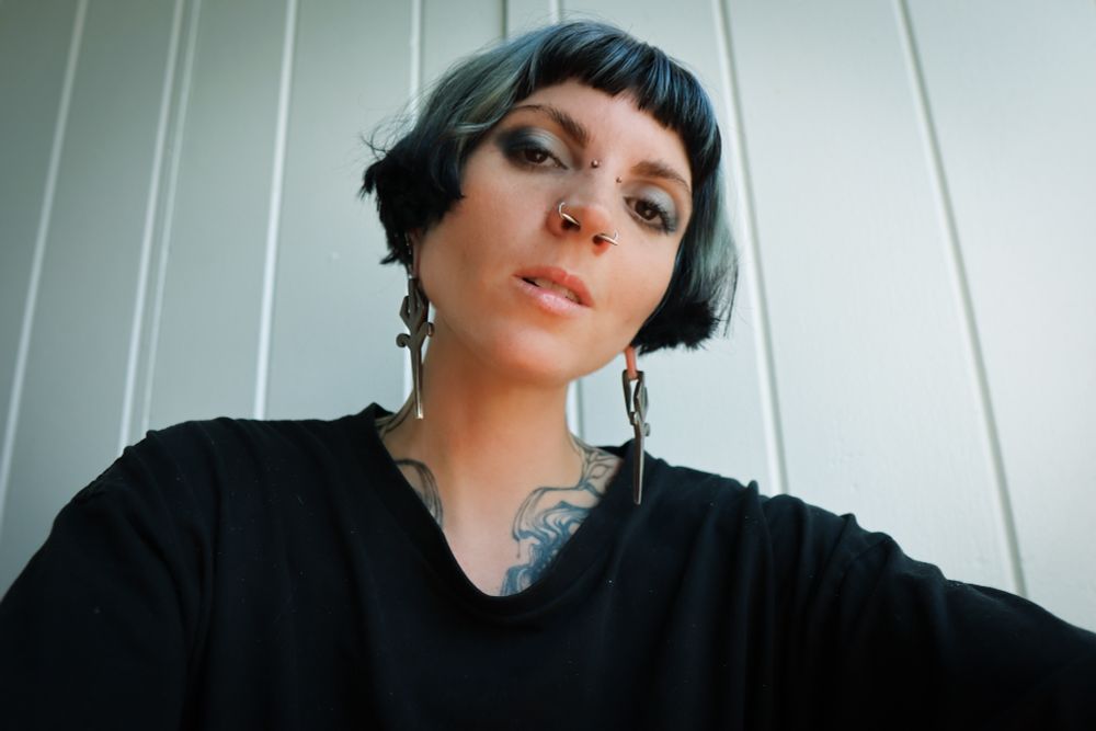 Cleo Kali sits in front of a plain panelled wall, positioned above the camera lens and making intense eye contact with the viewer. Her expression is calm, with only a flicker of wickedness in the way she holds her body, open and angled slightly, casually asserting a dominance already inherent. She wears a plain black t-shirt and solid silver ear weights, and her makeup is coordinated with her black and silver bob. A compelling liquid-drip chest tattoo is just visible, exposed where the collar of her t-shirt falls away.