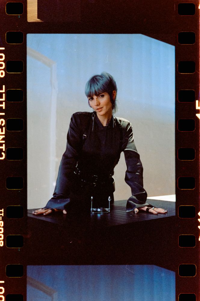Cleo Kali, captured on film, leans assertively over a desk with her palms flat and fingers outstretched, a steel CBT device seductively placed in front of her. Her eyes hold the camera as if directly into your own, daring you to approach.