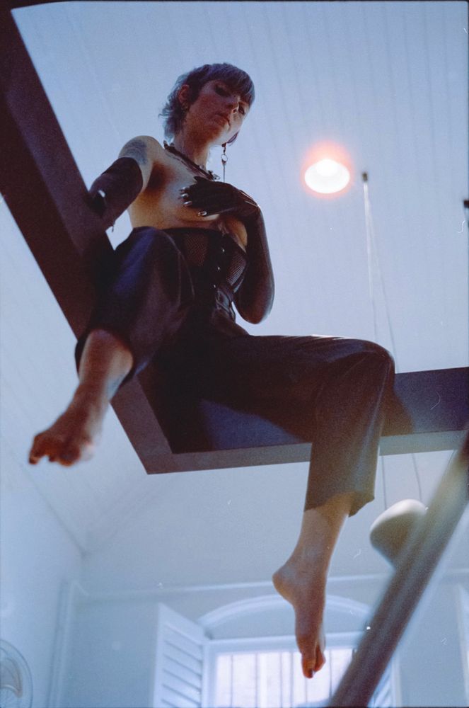 Cleo sits atop an orthogonal steel beam, topless and dressed in leather pants, a mesh corset, and elbow length leather gloves. Though her outfit is aesthetically arresting, her soft-focused bare feet are closest to the camera, commanding the viewer's gaze before all else. Once the viewer's eyes settle on her expression, they find Cleo staring expectantly down the lens, as if to dare a comment from whomever stares back.