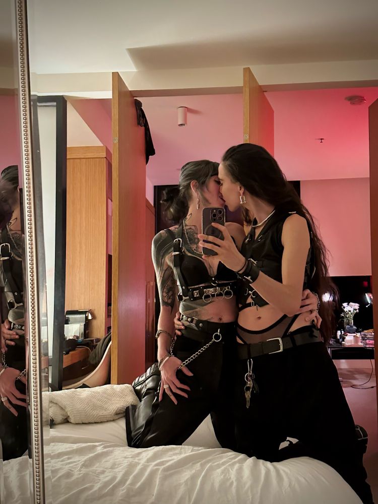 Cleo and Nyx kneel on a bed, capturing a passionate kiss in the mirror mid-pose. They are both adorned in layers of leather, silver steel and mesh, appropriately - and alluringly - dressed for a kinky queer play party.