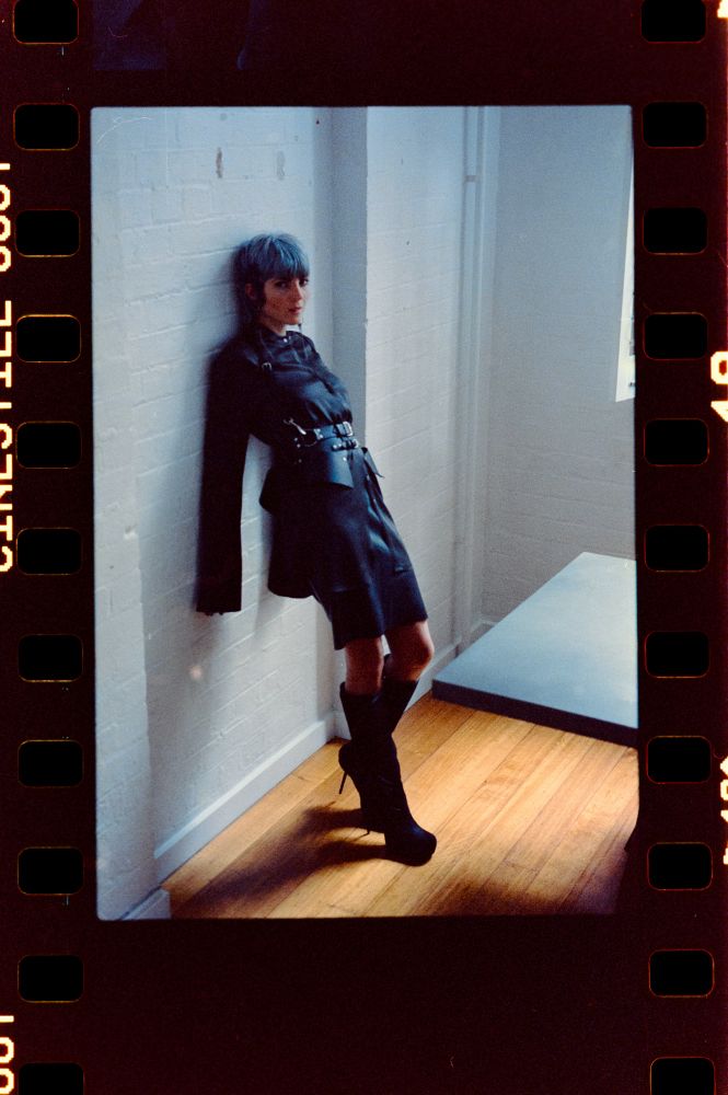 Cleo Kali, adorned in charcoal silk, black latex and leather accessories, leans against a white brick wall in the afternoon sunlight. Her expression suggests a weighted patience, neither expectant nor detached. What’s your next move?