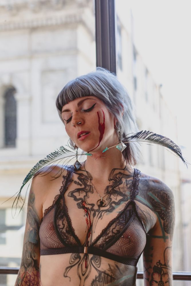Mistrix Cleo Kali - a heavily tattooed creature with short, choppy silver hair - poses in front of a floor to ceiling window that looks out into Naarm (Melbourne)’s sandstone CBD. Two rivulets of bright blood trickle from her right cheekbone down her chin and drip onto her chest, finding course through her cleavage. Two needles are crossed through her neck, each with a peacock feather extruding from the cap, orthogonally framing her face. She wears a serene expression, her eyes closed lightly and lips parted slightly. 