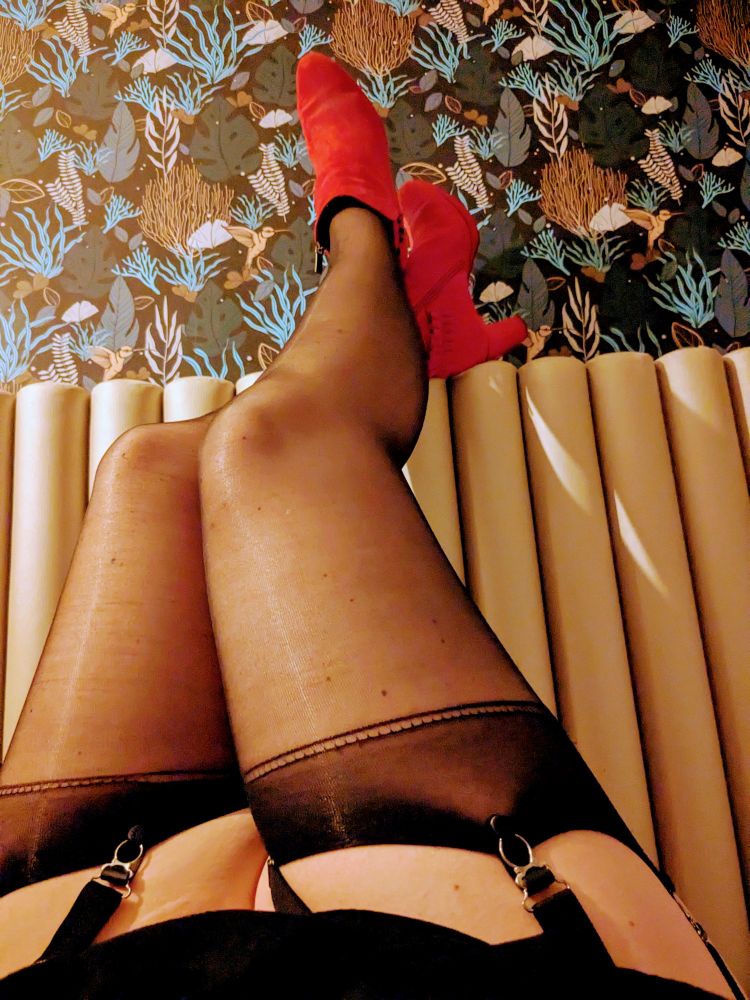 Beine an die Kopfwand eines Bettes gelehnt. Die Beine sind in Strapse gehüllt. Die Füße verschwinden in roten Stiefeletten.